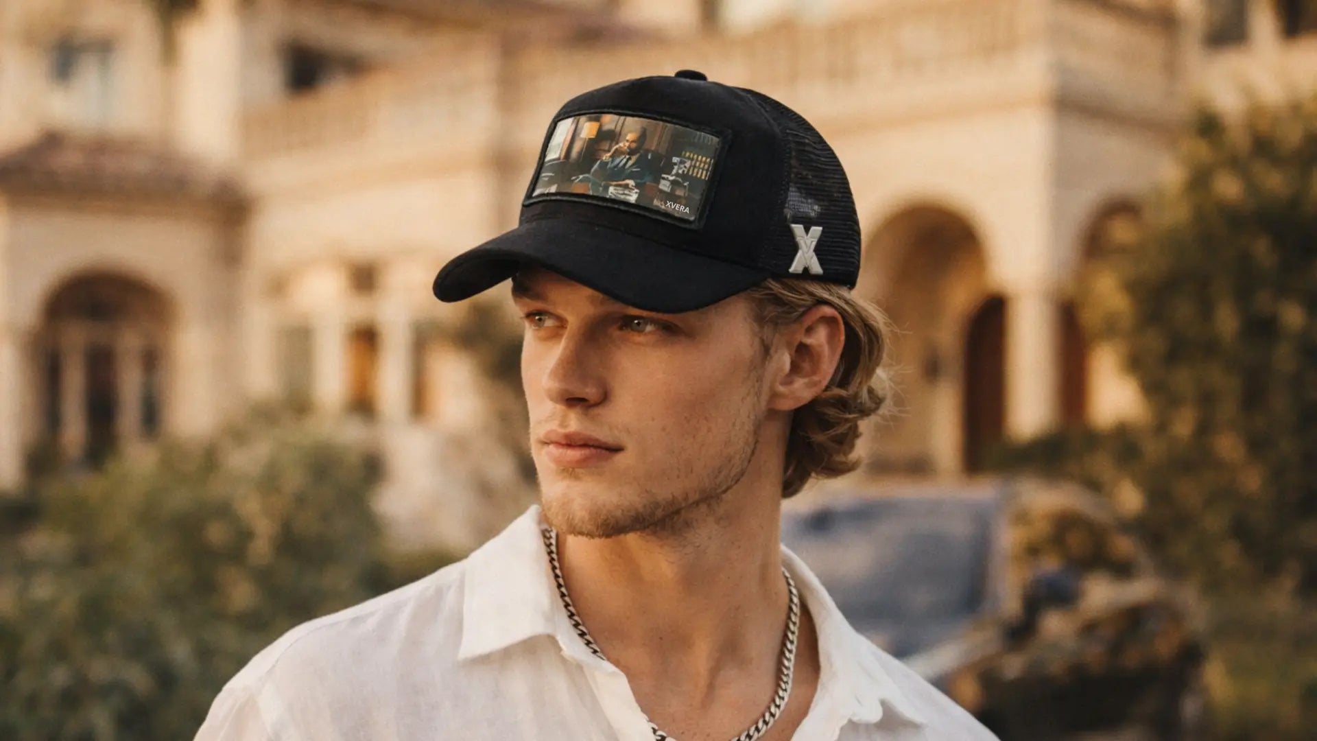Ung svensk man bär svart velvet trucker‑keps med metalliskt X-V‑emblem framför medelhavsvilla i gyllene solljus, lyxigt modefoto.