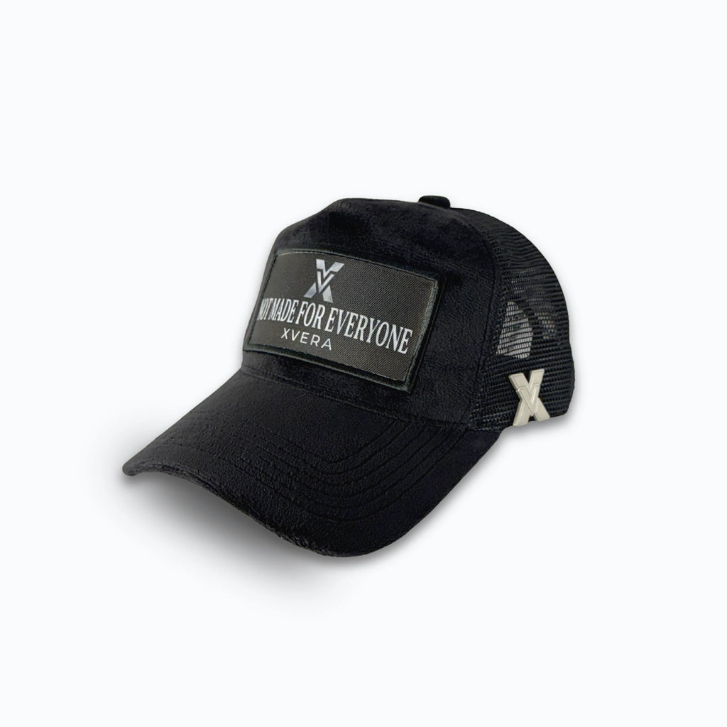 Svart keps i sammet från sidan, som visar passformen, den svarta nätbaksidan med X-logotyp i metall och patchen NOT MADE FOR EVERYONE. Unisex truckerkeps från XVERA.